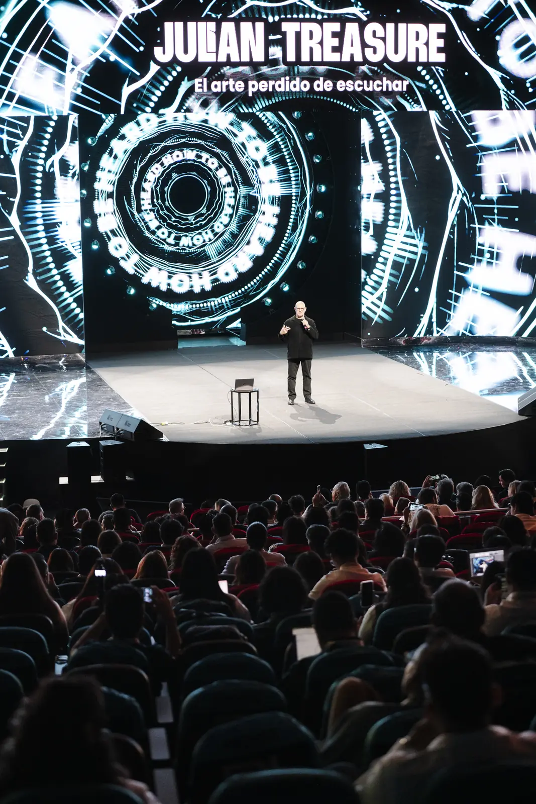 Julian Treasure on a large stage beneath a screen bearing his name, facing a full auditorium
