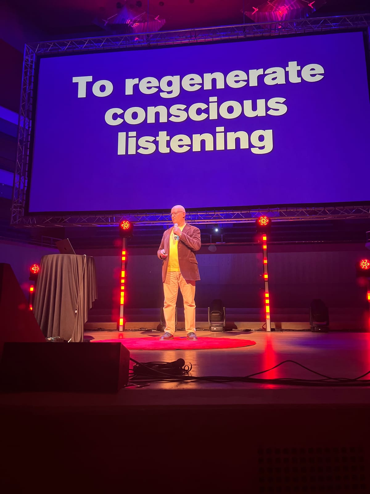 Julian Treasure on stage at TEDxManchester beneath a screen reading To regenerate conscious listening
