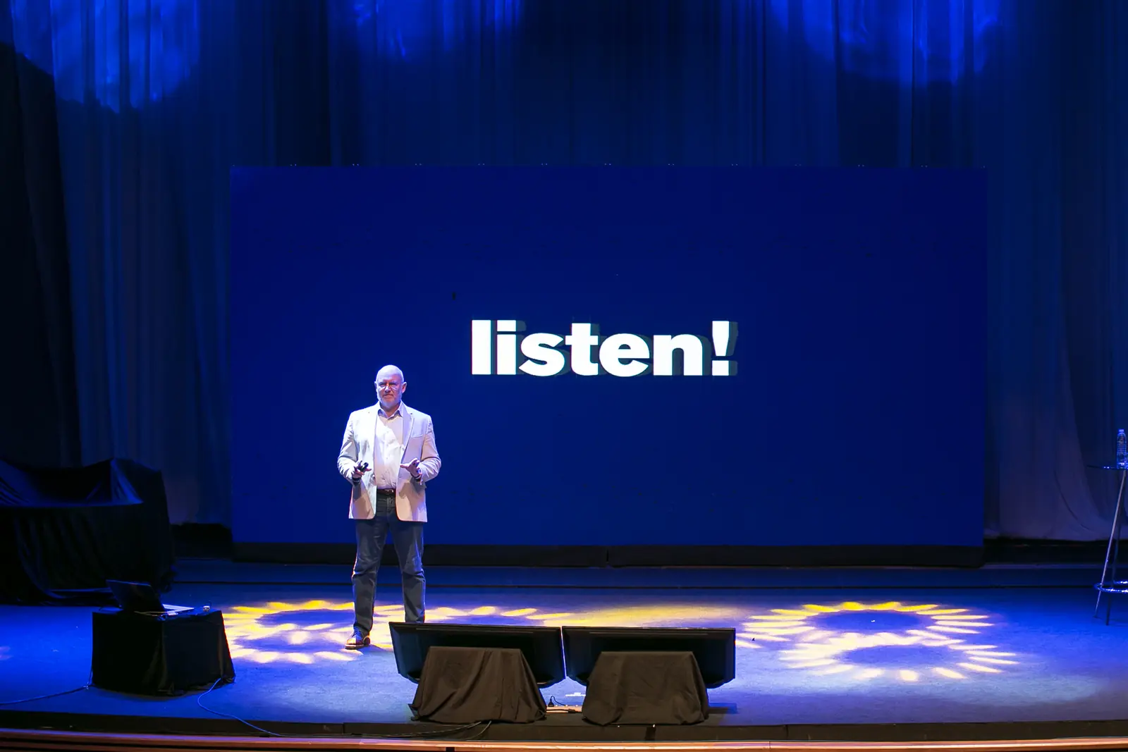 Julian Treasure on stage in front of a large blue screen reading listen!