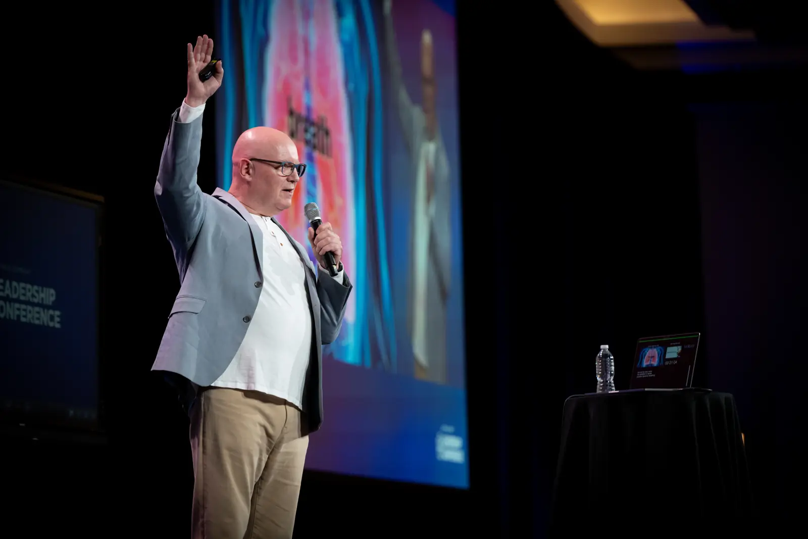 Julian Treasure on stage with a hand raised, in front of a large screen