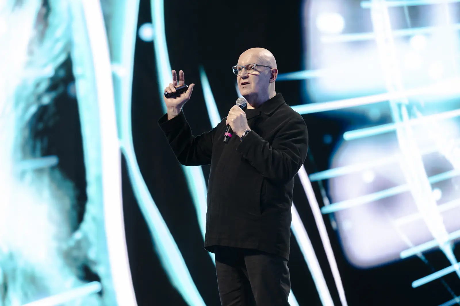Julian Treasure speaking into a handheld microphone, one hand raised, against a vivid screen