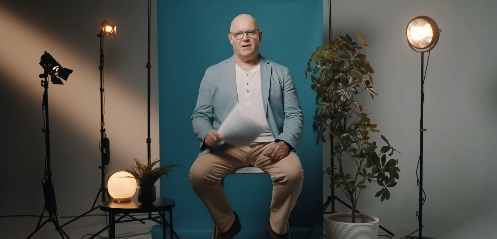 Julian Treasure seated on a stool in a film studio, with lights and a plant either side