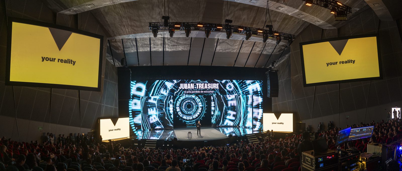 A panoramic view of a packed auditorium, with Julian Treasure on stage beneath a screen bearing his name and yellow side screens reading your reality