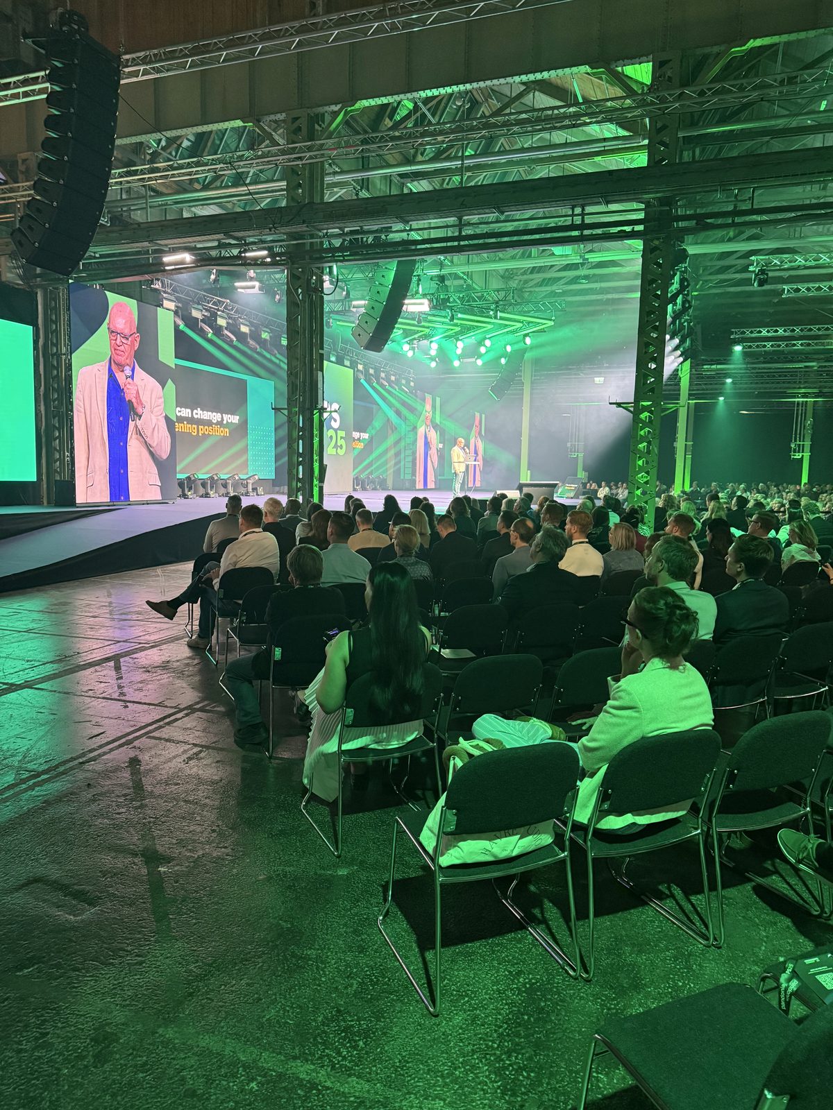 A large hall lit green, the audience in rows facing a distant stage, with Julian Treasure on the big screens