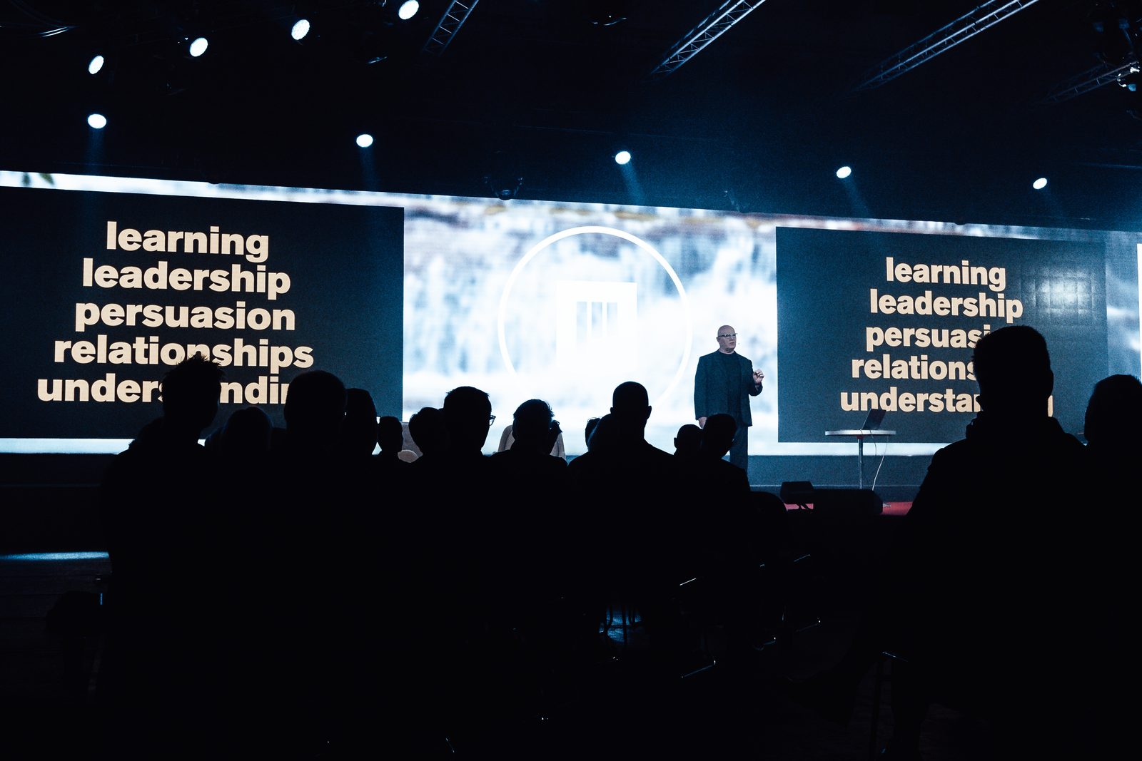 Julian Treasure on stage between two screens reading learning, leadership, persuasion, relationships, understanding, the audience in silhouette