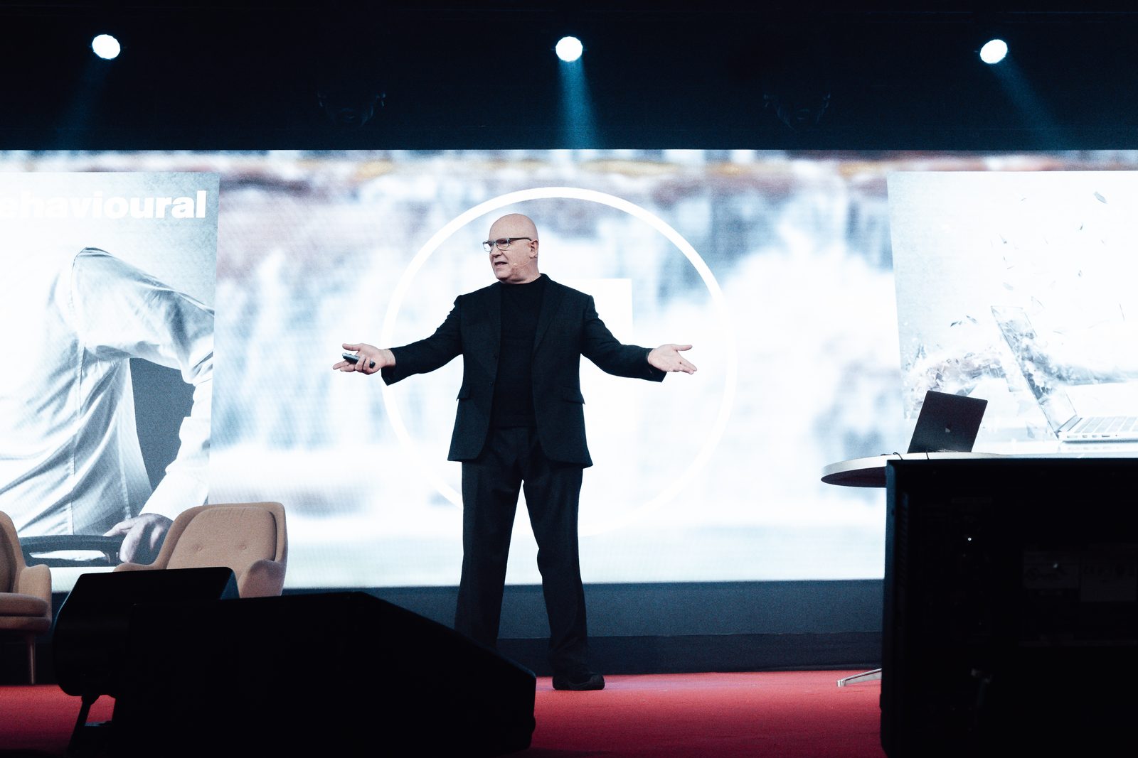 Julian Treasure in black with arms open on a red-floored stage, in front of a wide screen