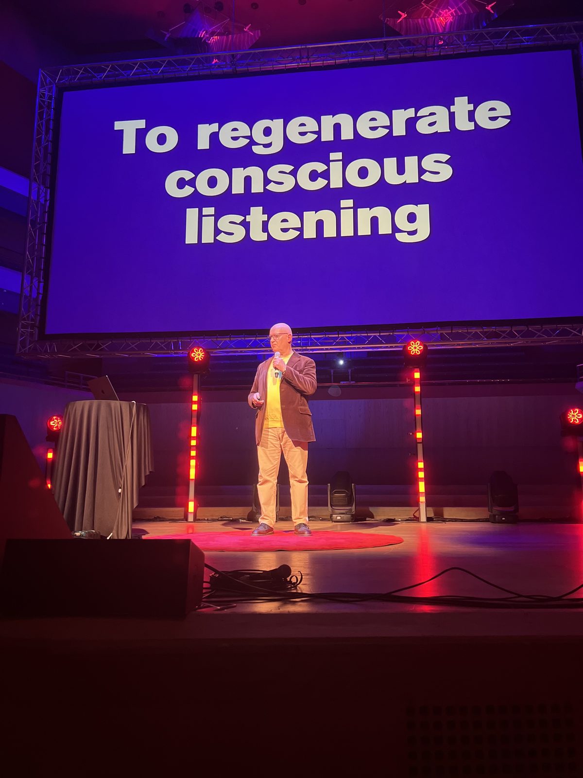 Julian Treasure on stage at TEDxManchester beneath a screen reading To regenerate conscious listening
